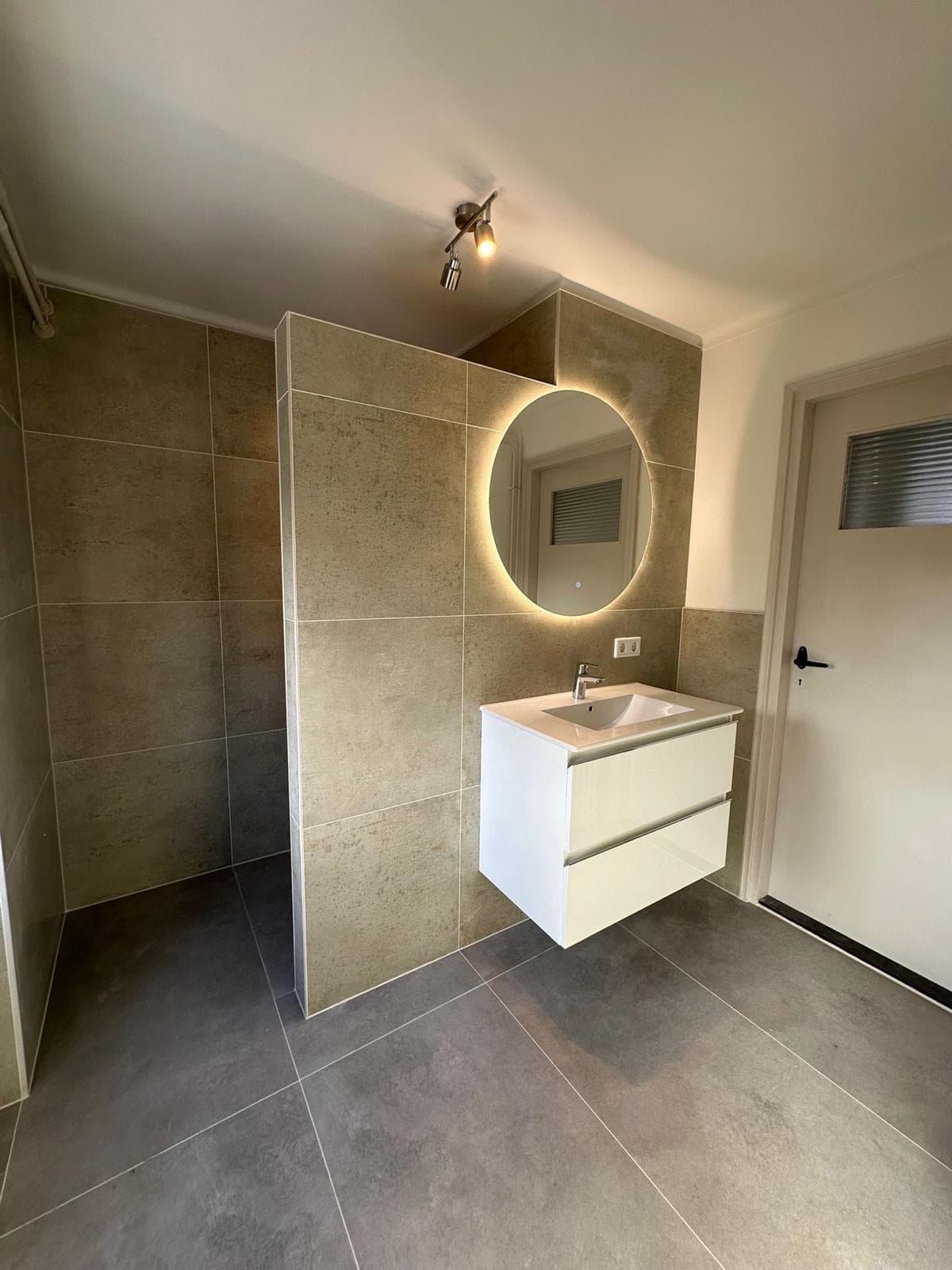Badkamer met inloopdouche, ronde verlichte spiegel, zwevend wit wastafelmeubel en grootformaattegels — Vesta Lux project in Zeeland